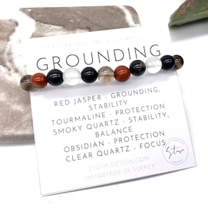 May include: A grounding bracelet featuring red jasper, tourmaline, smoky quartz, obsidian, and clear quartz beads. The bracelet is displayed on a white card with the text "GROUNDING" and the properties of each stone.