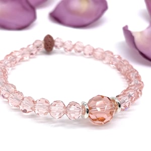 Könnte beinhalten: Ein rosafarbenes Kristall-Armband mit einer größeren rosafarbenen Kristallperle in der Mitte. Das Armband besteht aus kleinen, facettierten Kristallperlen und einer braunen Lavaperle.