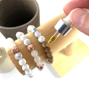 May include: Three bracelets on a wooden display stand. The bracelets are made of white beads, brown wood beads, and lava beads with rose gold accents. A dropper bottle of essential oil is being used to add oil to the lava beads.