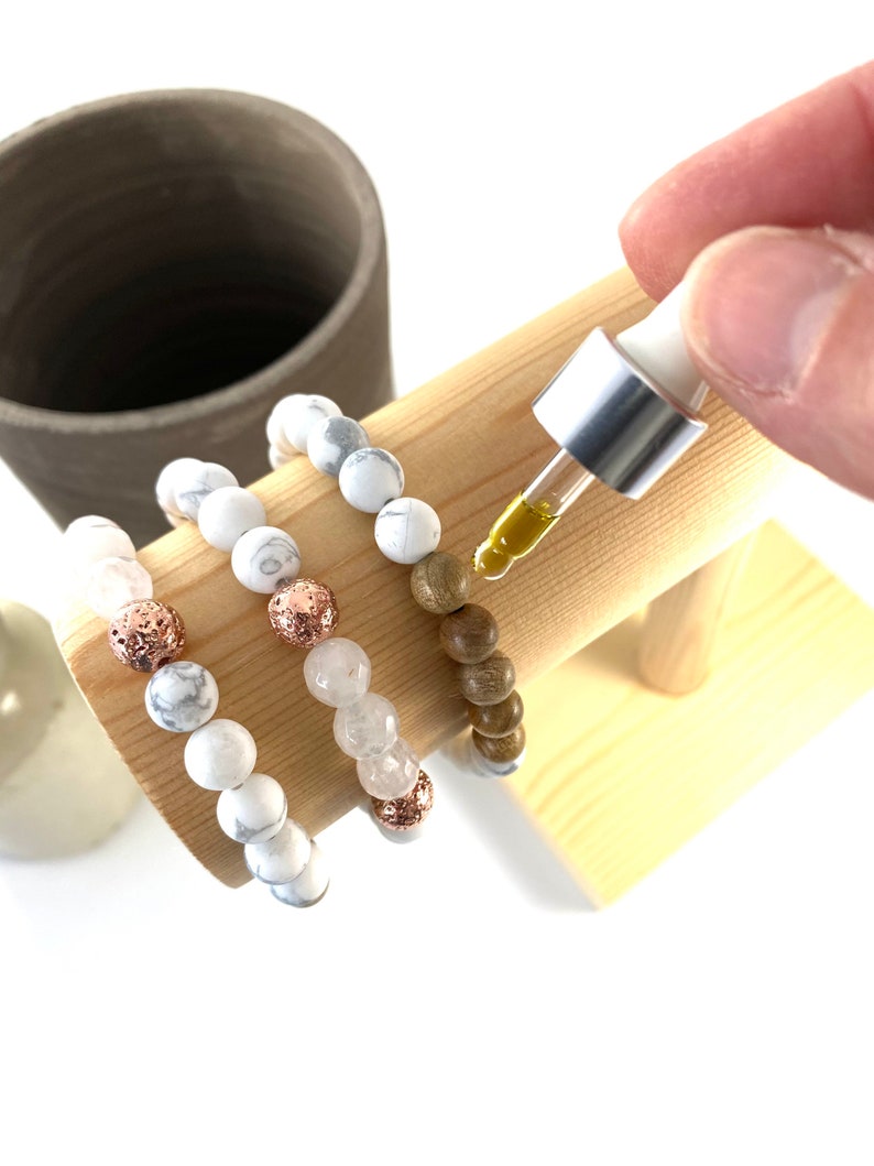 May include: Three bracelets on a wooden display stand. The bracelets are made of white beads, brown wood beads, and lava beads with rose gold accents. A dropper bottle of essential oil is being used to add oil to the lava beads.