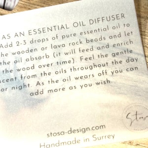 May include: White card with black text explaining how to use an essential oil diffuser. The card says "As an essential oil diffuser, add 2-3 drops of pure essential oil to the wooden or lava rock beads and let the oil absorb (it will feed and enrich the wood over time). Feel the gentle scent from the oils throughout the day or night. As the oil wears off you can add more as you wish." The card also has the text "stosa-design.com Handmade in Surrey" and a logo that says "Stoga".