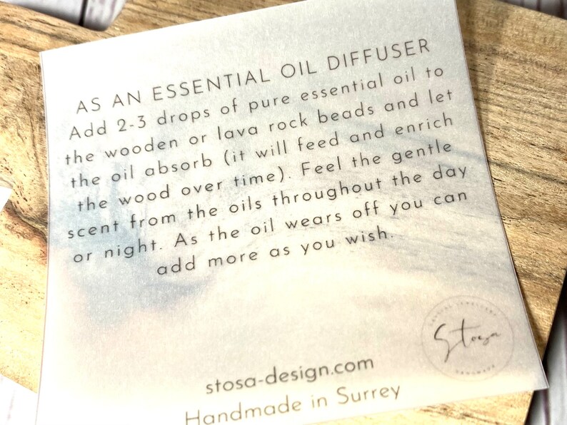 May include: White card with black text explaining how to use an essential oil diffuser. The card says "As an essential oil diffuser, add 2-3 drops of pure essential oil to the wooden or lava rock beads and let the oil absorb (it will feed and enrich the wood over time). Feel the gentle scent from the oils throughout the day or night. As the oil wears off you can add more as you wish." The card also has the text "stosa-design.com Handmade in Surrey" and a logo that says "Stoga".