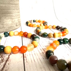 May include: Three bracelets made of orange, green, and brown beads. The bracelets are made of natural stones and are stacked on a white wooden surface.