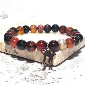 Könnte beinhalten: Ein Armband mit bunten Perlen in Rot-, Braun-, Schwarz- und Goldtönen. Ein silberner Katzenanhänger hängt am Armband. Das Armband wird auf einer hellen, strukturierten Oberfläche präsentiert.