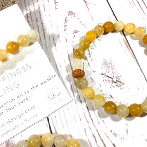 Pu&ograve; includere: Un braccialetto di perline gialle e bianche con un ciondolo di legno. Il braccialetto &egrave; su una superficie di legno bianco con un biglietto che dice "Happiness - Healing" e "stosa-design.com".