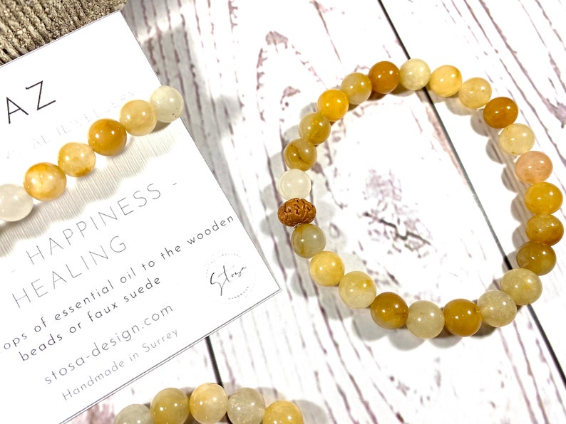 Pu&ograve; includere: Un braccialetto di perline gialle e bianche con un ciondolo di legno. Il braccialetto &egrave; su una superficie di legno bianco con un biglietto che dice "Happiness - Healing" e "stosa-design.com".
