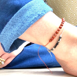 Op de afbeelding: Twee kralenanklets om de enkel van een persoon. De bovenste anklet is gemaakt van rode kralen en de onderste anklet is gemaakt van zwarte kralen.