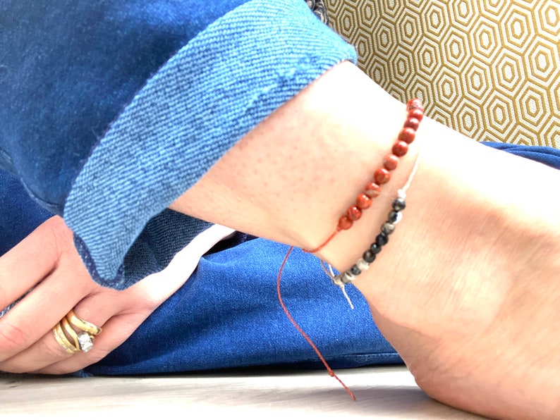 Op de afbeelding: Twee kralenanklets om de enkel van een persoon. De bovenste anklet is gemaakt van rode kralen en de onderste anklet is gemaakt van zwarte kralen.