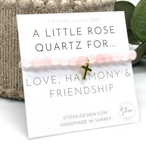 Puede incluir: Un brazalete de cuarzo rosa rosa con un dije de cruz dorada. El brazalete está sobre una tarjeta blanca con el texto "A Little Rose Quartz For... Love, Harmony & Friendship" y "STOSA-DESIGN.COM HANDMADE IN SURREY".