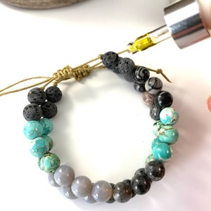 May include: A beaded bracelet with a mix of gray, black, and turquoise stones. A dropper bottle with yellow liquid is being used to add essential oils to the lava rock beads.