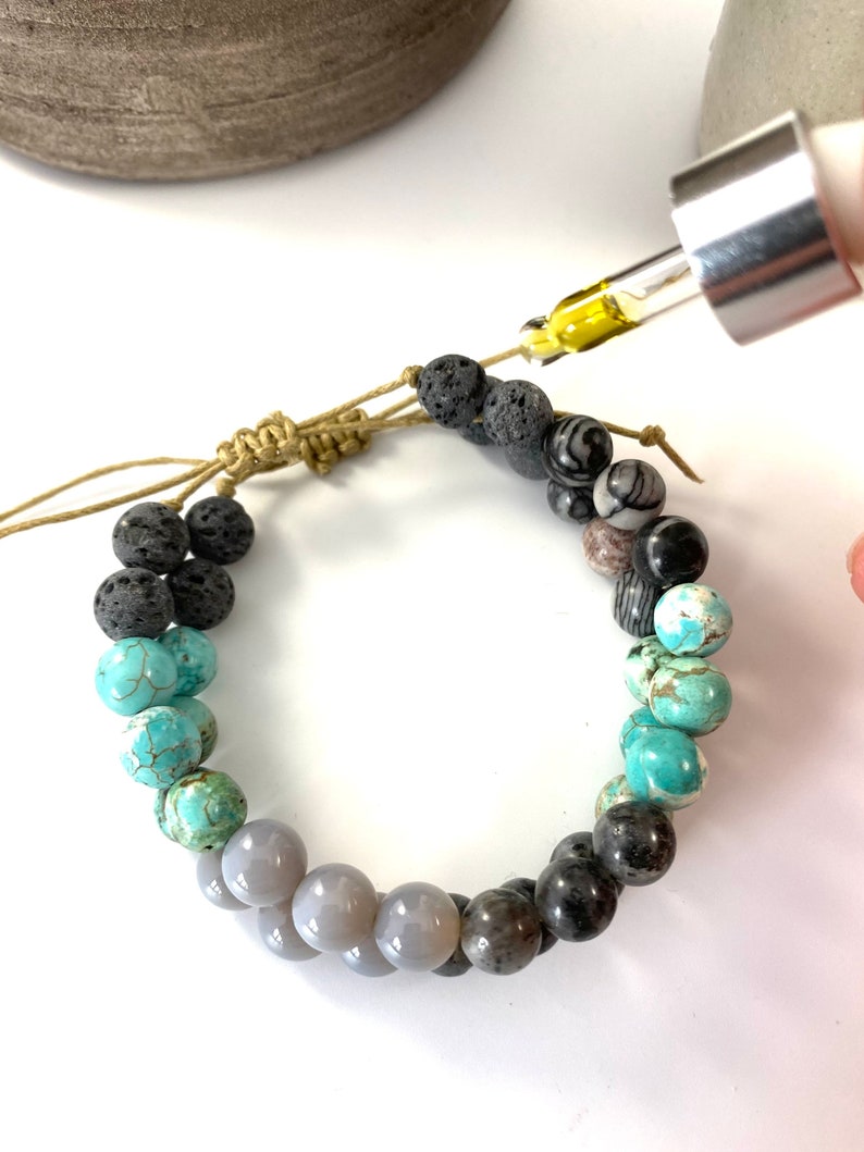 May include: A beaded bracelet with a mix of gray, black, and turquoise stones. A dropper bottle with yellow liquid is being used to add essential oils to the lava rock beads.