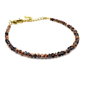 Op de afbeelding: Een delicate armband gemaakt met kleine, gefacetteerde glazen kralen in koper- en paarse tinten. De armband wordt vastgemaakt met een gouden sluiting.