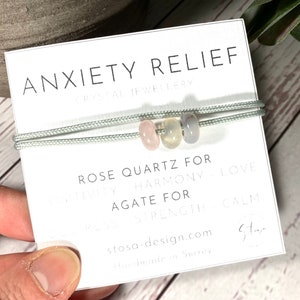 Puede incluir: Un brazalete de cuerda gris con tres cuentas: una cuenta de cuarzo rosa, una cuenta de ágata transparente y una cuenta de ágata gris claro. El brazalete está sobre una tarjeta blanca con el texto "ANXIETY RELIEF", "CRYSTAL JEWELLERY", "ROSE QUARTZ FOR", "HARMONY LOVE", "AGATE FOR", "STRENGTH CALM", "stosa-design.com" y "Handmade in Surrey".