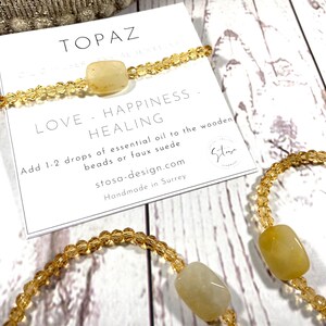 May include: Two topaz crystal bracelets with gold beads. The bracelets are on a white wooden surface with a card that reads "Topaz Crystal Jewelry Love - Happiness - Healing Add 1-2 drops of essential oil to the wooden beads or faux suede stosa-design.com Handmade in Surrey Stay"