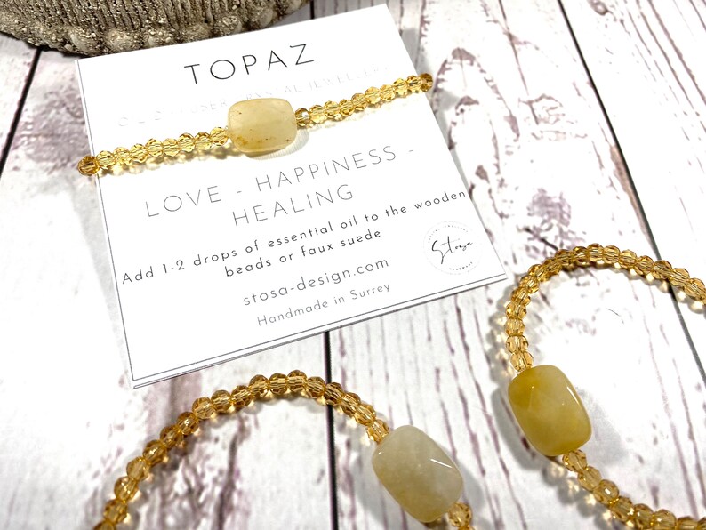 May include: Two topaz crystal bracelets with gold beads. The bracelets are on a white wooden surface with a card that reads "Topaz Crystal Jewelry Love - Happiness - Healing Add 1-2 drops of essential oil to the wooden beads or faux suede stosa-design.com Handmade in Surrey Stay"