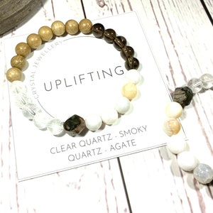 May include: A bracelet made with clear quartz, smoky quartz, and agate beads. The bracelet is on a white card with the text "CRYSTAL JEWELLERY - UPLIFTING - CLEAR QUARTZ - SMOKY QUARTZ - AGATE"