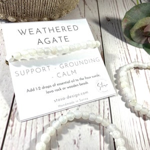 Puede incluir: Dos pulseras de cuentas de ágata blanca sobre una superficie de madera blanca. Las pulseras están apiladas una encima de la otra. Una tarjeta blanca con el texto "WEATHERED AGATE SUPPORT - GROUNDING - CALM Add 1-2 drops of essential oil to the faux suede, lava rock or wooden beads stosa-design.com Handmade in Surrey" está detrás de las pulseras.