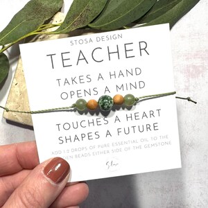 Puede incluir: Un brazalete de cuentas verdes y marrones con el texto "STOSA DESIGN TEACHER TAKES A HAND OPENS A MIND TOUCHES A HEART SHAPES A FUTURE ADD 1 2 DROPS OF PURE ESSENTIAL OIL TO THE DEN BEADS EITHER SIDE OF THE GEMSTONE Star" en una tarjeta blanca.