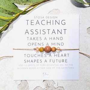 May include: A white card with the text "STOSA DESIGN TEACHING ASSISTANT TAKES A HAND OPENS A MIND TOUCHES A HEART SHAPES A FUTURE ADD 1-2 DROPS OF PURE ESSENTIAL OIL TO THE WOODEN BEADS EITHER SIDE OF THE GEMSTONE" and a brown string bracelet with wooden beads and a gemstone.