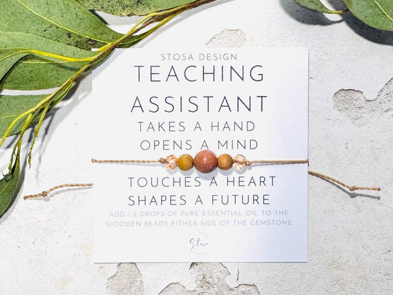 May include: A white card with the text "STOSA DESIGN TEACHING ASSISTANT TAKES A HAND OPENS A MIND TOUCHES A HEART SHAPES A FUTURE ADD 1-2 DROPS OF PURE ESSENTIAL OIL TO THE WOODEN BEADS EITHER SIDE OF THE GEMSTONE" and a brown string bracelet with wooden beads and a gemstone.
