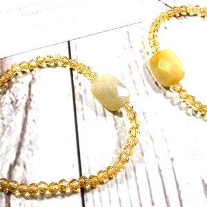 May include: Two bracelets with gold beads and a light yellow gemstone. The bracelets are on a white wooden surface.