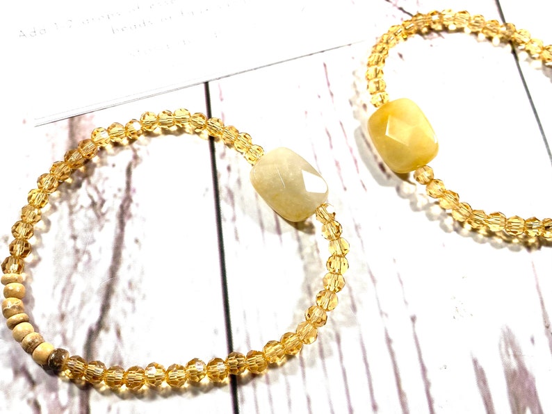May include: Two bracelets with gold beads and a light yellow gemstone. The bracelets are on a white wooden surface.