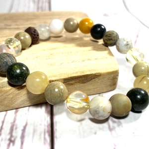 May include: A bracelet made of various natural stones, including brown, white, yellow, and green stones. The bracelet is on a wooden surface.