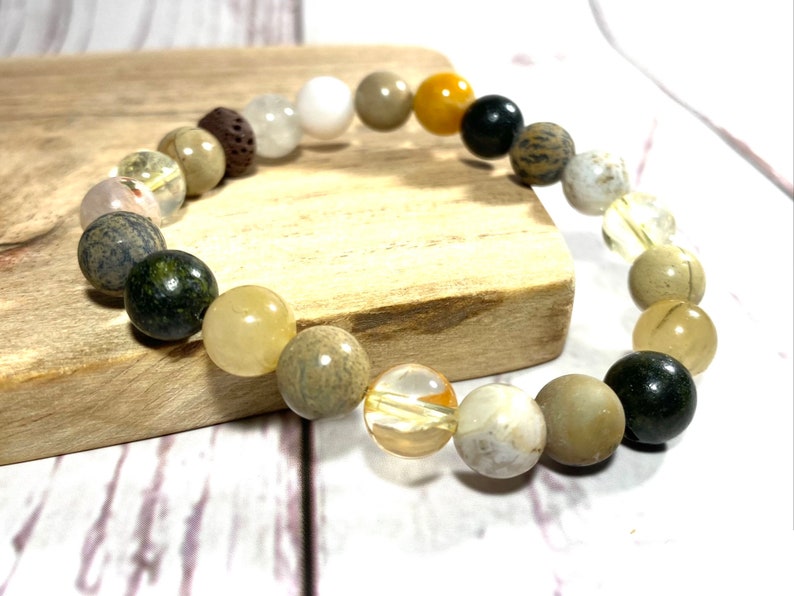 May include: A bracelet made of various natural stones, including brown, white, yellow, and green stones. The bracelet is on a wooden surface.