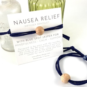 May include: A navy blue diffuser bracelet with a wooden bead and a blue stone. The bracelet is designed to be worn with the bead over the P6 acupressure point on the inside of the wrist. The card reads "Nausea Relief Diffuser Bracelet with Blue Spot Jasper for Mindfulness Calm Support. Designed to be worn with the wooden bead over the P6 acupressure point on the inside of the wrist. You can also add essential oil to the bead and faux suede for extra support. www.albion-design.com Handmade in Surrey."