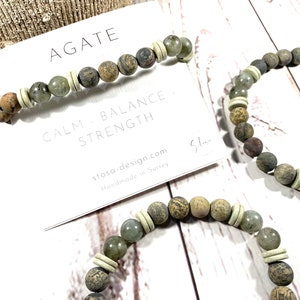 May include: Two agate stone bracelets with white spacer beads. The bracelets are on a white surface with a card that reads "Agate, Calm - Balance - Strength, stosa-design.com, Handmade in Surrey".