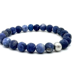 Könnte beinhalten: Ein Armband mit dunkelblauen und silbernen Perlen. Das Armband besteht aus runden, mattierten Perlen in verschiedenen Blautönen, mit einer einzelnen silbernen Perle. Auf weißem Hintergrund.