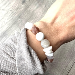 May include: A bracelet with a mix of white, grey, and pink gemstones. The bracelet is worn on a wrist, with a grey long-sleeved top visible. The gemstones are various shapes and sizes, creating a unique accessory.