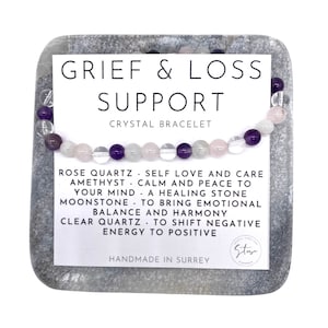 May include: A crystal bracelet featuring rose quartz, amethyst, moonstone, and clear quartz beads. The bracelet is presented on a grey stone with a card that reads "GRIEF & LOSS SUPPORT CRYSTAL BRACELET". The card lists crystal properties.