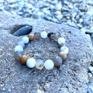 May include: A bracelet made of white, gray, and brown beads. The beads are round and have a smooth, polished finish. The bracelet is on a gray, textured surface.