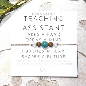 May include: A white card with the text "STOSA DESIGN TEACHING ASSISTANT TAKES A HAND OPENS A MIND TOUCHES A HEART SHAPES A FUTURE ADD 12 DROPS OF PURE ESSENTIAL OIL TO THE WOODEN BEADS EITHER SIDE OF THE GEMSTONE" and a signature. A string with wooden beads and a turquoise gemstone is wrapped around the card.