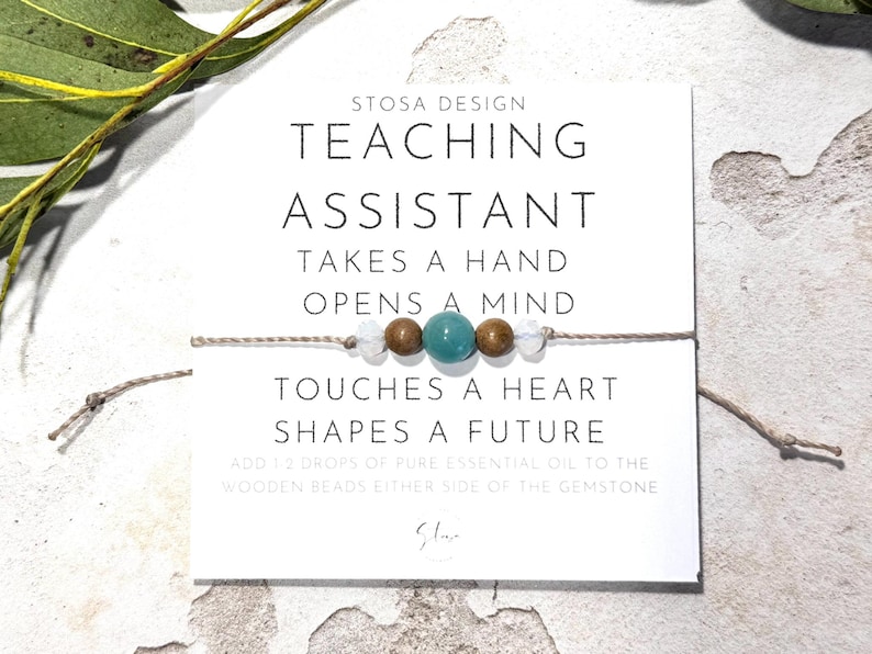 May include: A white card with the text "STOSA DESIGN TEACHING ASSISTANT TAKES A HAND OPENS A MIND TOUCHES A HEART SHAPES A FUTURE ADD 12 DROPS OF PURE ESSENTIAL OIL TO THE WOODEN BEADS EITHER SIDE OF THE GEMSTONE" and a signature. A string with wooden beads and a turquoise gemstone is wrapped around the card.