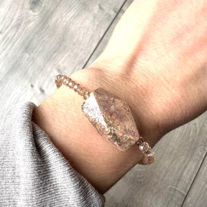 May include: A gemstone bracelet with a large, irregularly shaped, light brown crystal as the focal point. The crystal is flanked by smaller, round, light brown beads. The bracelet is worn on a wrist, with a grey sleeve visible.