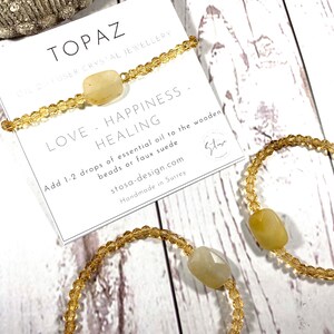 May include: Three gold beaded bracelets with a topaz crystal charm. The bracelets are on a white wooden surface. The card reads "Topaz Diffuser Crystal Jewellery Love - Happiness - Healing Add 1-2 drops of essential oil to the wooden beads or faux suede stosa-design.com Handmade in Surrey"