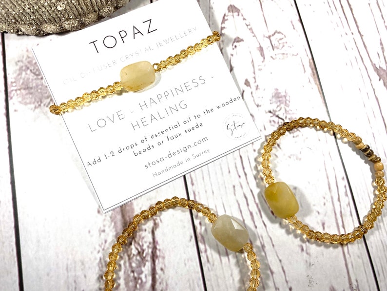 May include: Three gold beaded bracelets with a topaz crystal charm. The bracelets are on a white wooden surface. The card reads "Topaz Diffuser Crystal Jewellery Love - Happiness - Healing Add 1-2 drops of essential oil to the wooden beads or faux suede stosa-design.com Handmade in Surrey"