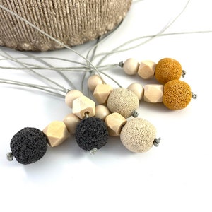 May include: A set of five necklaces with different colored beads. The necklaces are made with natural materials, including wood, lava rock, and beige stone. The necklaces are all strung on a gray cord.