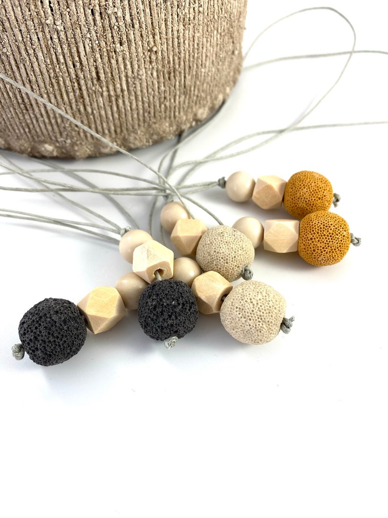 May include: A set of five necklaces with different colored beads. The necklaces are made with natural materials, including wood, lava rock, and beige stone. The necklaces are all strung on a gray cord.
