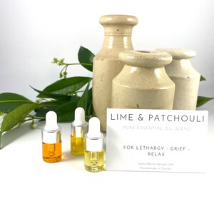 May include: Three small bottles of essential oil blend with white dropper tops and a yellow liquid. The bottles are arranged on a white surface. A white card with the text "LIME & PATCHOULI PURE ESSENTIAL OIL BLEND FOR LETHARGY GRIEF RELAX www.albion-design.com Handmade in Surrey" is in the background.