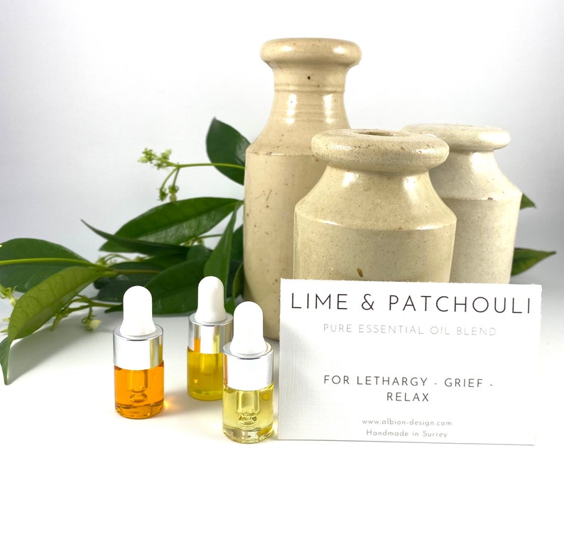 May include: Three small bottles of essential oil blend with white dropper tops and a yellow liquid. The bottles are arranged on a white surface. A white card with the text "LIME & PATCHOULI PURE ESSENTIAL OIL BLEND FOR LETHARGY GRIEF RELAX www.albion-design.com Handmade in Surrey" is in the background.