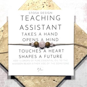 May include: A white card with the text "STOSA DESIGN TEACHING ASSISTANT TAKES A HAND OPENS A MIND TOUCHES A HEART SHAPES A FUTURE ADD 1 2 DROPS OF PURE ESSENTIAL OIL TO THE WOODEN BEADS EITHER SIDE OF THE GEMSTONE" and a small pink gemstone bracelet with brown wooden beads.