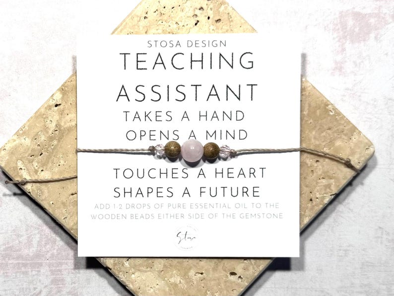 May include: A white card with the text "STOSA DESIGN TEACHING ASSISTANT TAKES A HAND OPENS A MIND TOUCHES A HEART SHAPES A FUTURE ADD 1 2 DROPS OF PURE ESSENTIAL OIL TO THE WOODEN BEADS EITHER SIDE OF THE GEMSTONE" and a small pink gemstone bracelet with brown wooden beads.