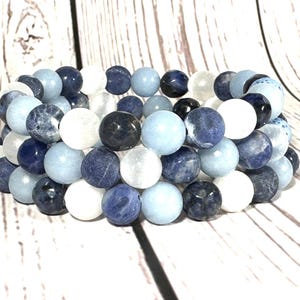May include: Three beaded bracelets, each made of round beads in white, light blue, and dark blue. The beads have a polished finish and are stacked. The background is a light wood grain. The bracelets are a stylish accessory.
