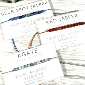 Op de afbeelding: Drie armbanden gemaakt met verschillende soorten edelstenen en vastgebonden met koord. De armbanden zijn gelabeld met het type steen en de bijbehorende voordelen: Blauwe vlekjaspis - Mindfulness, Moed, Kalmte; Rode jaspis - Stressvermindering, Evenwicht, Aarding; Agate - Kalmte, Evenwicht, Kracht.