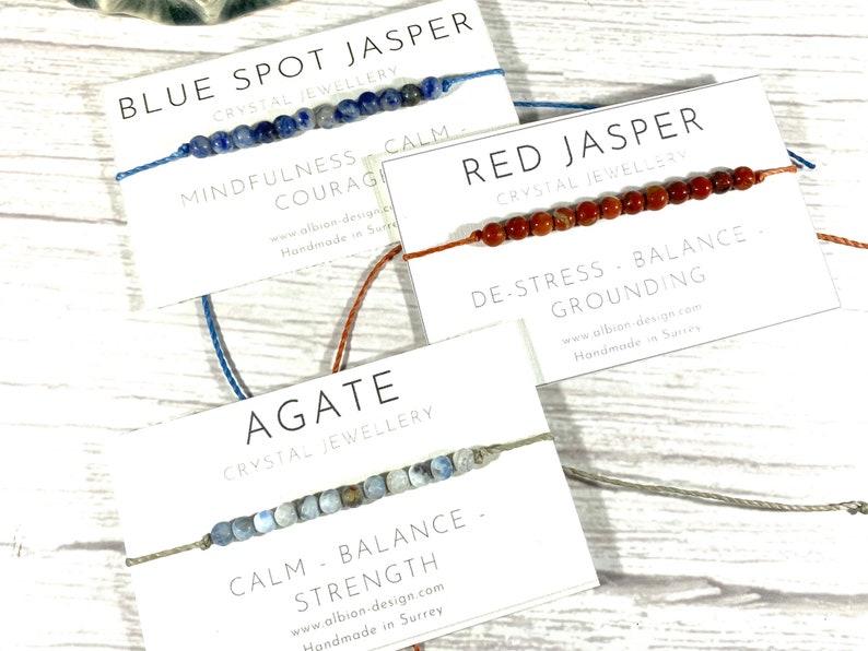Op de afbeelding: Drie armbanden gemaakt met verschillende soorten edelstenen en vastgebonden met koord. De armbanden zijn gelabeld met het type steen en de bijbehorende voordelen: Blauwe vlekjaspis - Mindfulness, Moed, Kalmte; Rode jaspis - Stressvermindering, Evenwicht, Aarding; Agate - Kalmte, Evenwicht, Kracht.