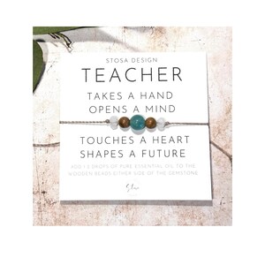Puede incluir: Una tarjeta blanca con el texto "STOSA DESIGN TEACHER TAKES A HAND OPENS A MIND TOUCHES A HEART SHAPES A FUTURE ADD 12 DROPS OF PURE ESSENTIAL OIL TO THE WOODEN BEADS EITHER SIDE OF THE GEMSTONE Star" y una pulsera de cuerda marr&oacute;n con cuentas blancas y marrones y una piedra preciosa turquesa.