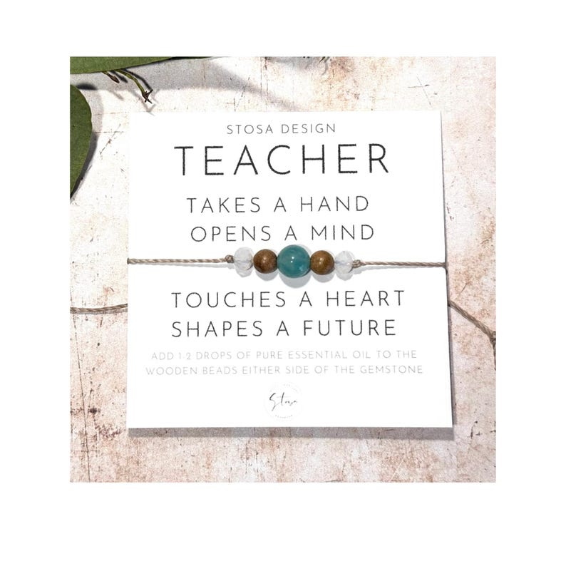 Puede incluir: Una tarjeta blanca con el texto "STOSA DESIGN TEACHER TAKES A HAND OPENS A MIND TOUCHES A HEART SHAPES A FUTURE ADD 12 DROPS OF PURE ESSENTIAL OIL TO THE WOODEN BEADS EITHER SIDE OF THE GEMSTONE Star" y una pulsera de cuerda marr&oacute;n con cuentas blancas y marrones y una piedra preciosa turquesa.